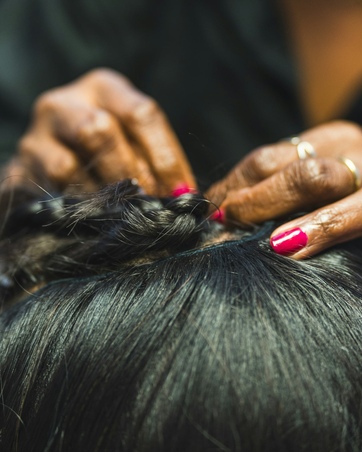 Hands braiding dark hair with pink nails.